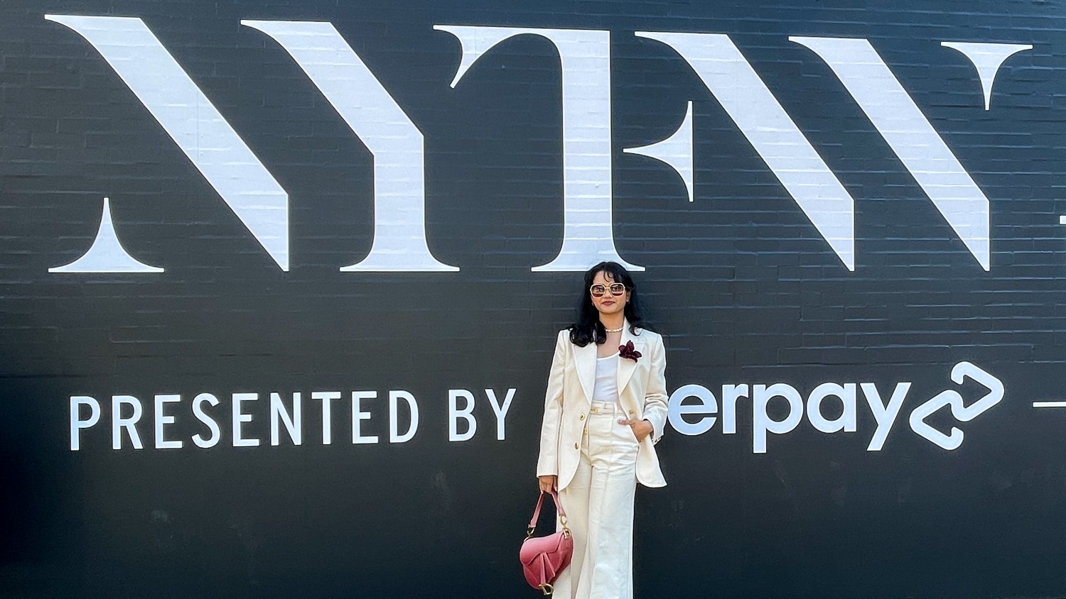 Photo of Ankitha Gurram in front of a black brick mural painted with the NYFW logo in white. Gurram wears an off-white suit.