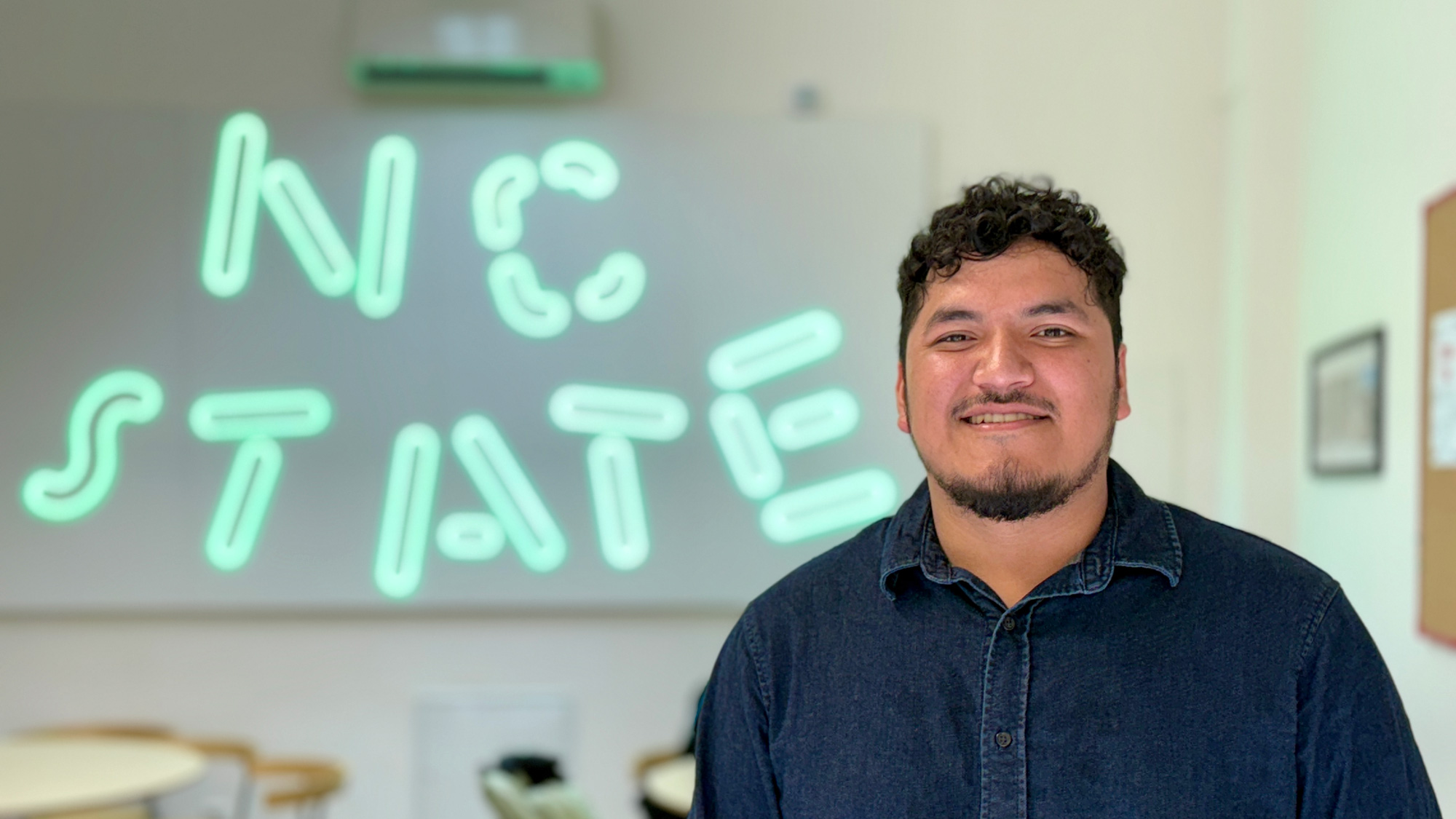 Angel Barron-Rueda smiling in front of "NC State" in neon
