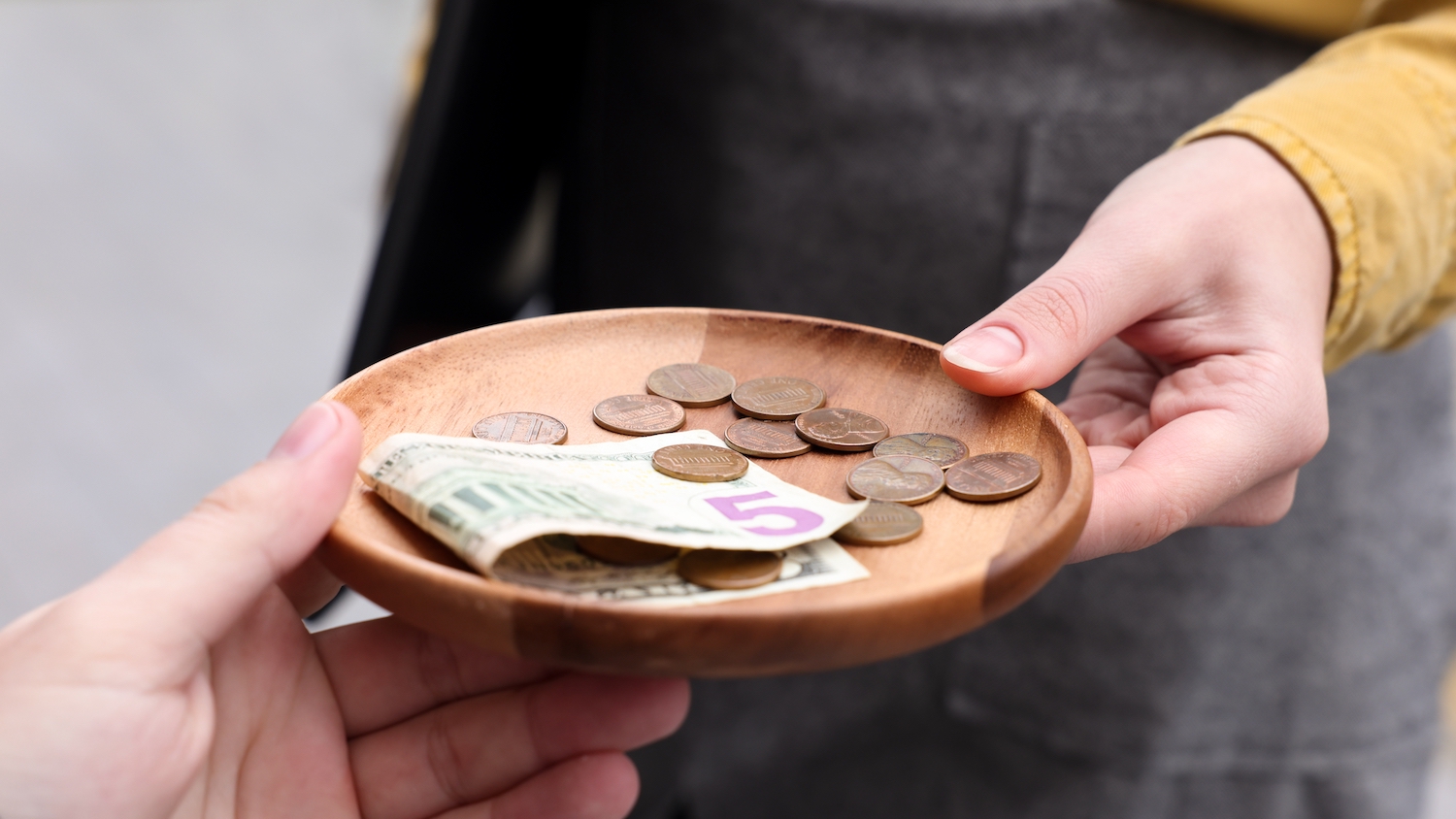 A hand passes a wooden saucer will bills and coins to another hand.