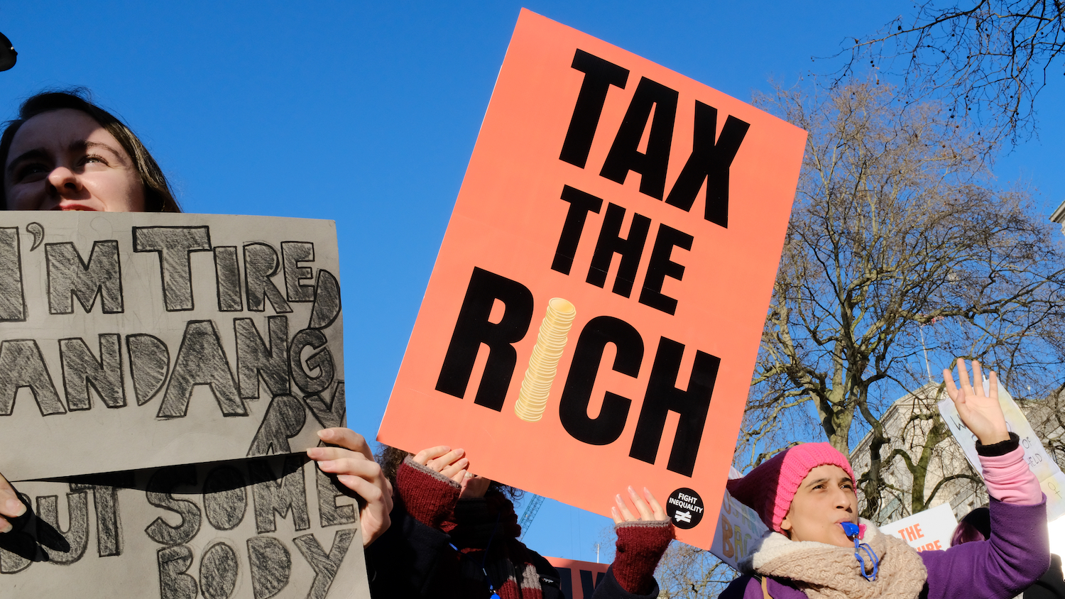 A woman in a crowd holds up an orange sign that reads "Tax the rich."