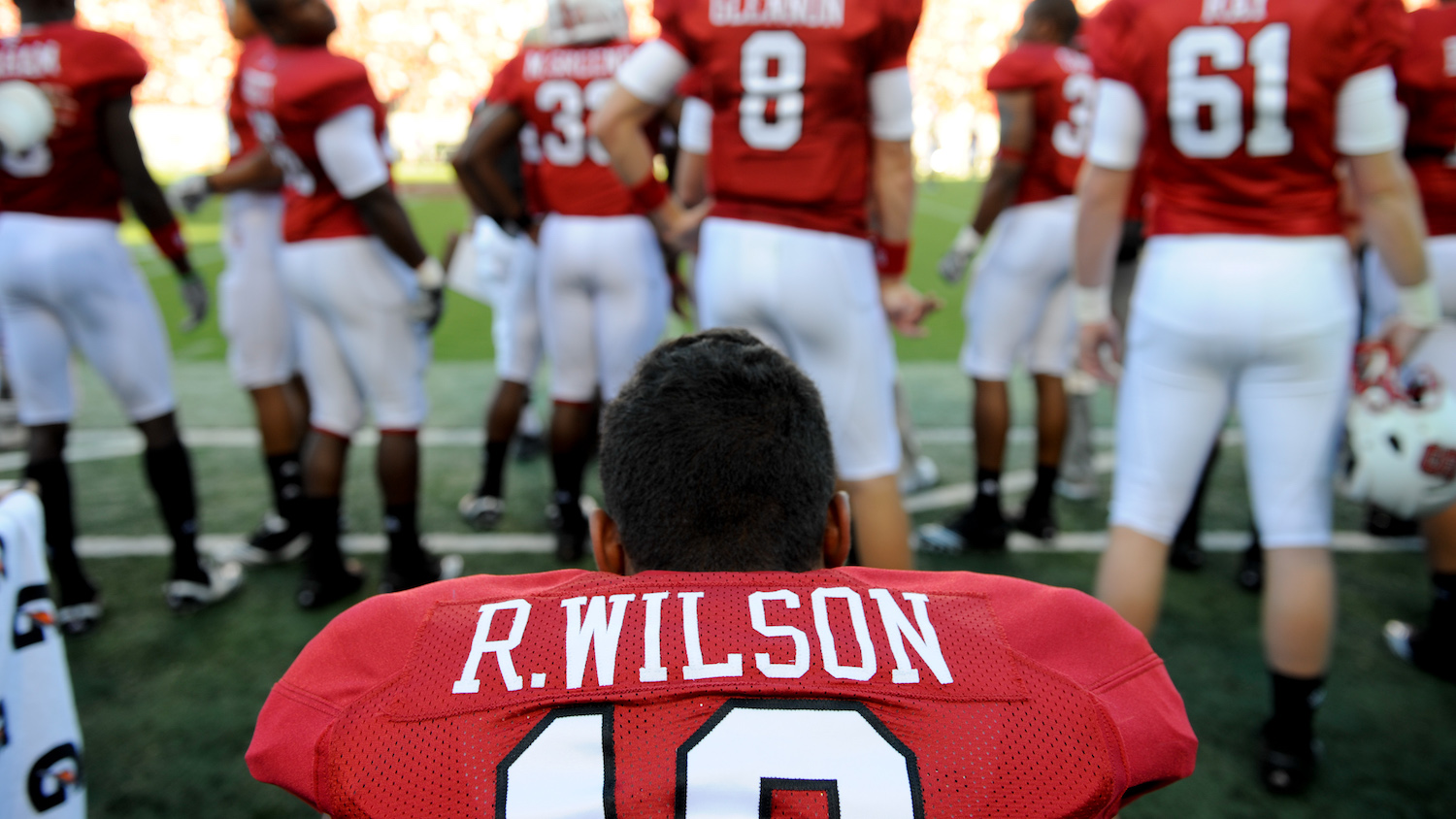 Football player Russell Wilson, back to the camera, sits on the bunch as other NC State football players stand on the sideline.