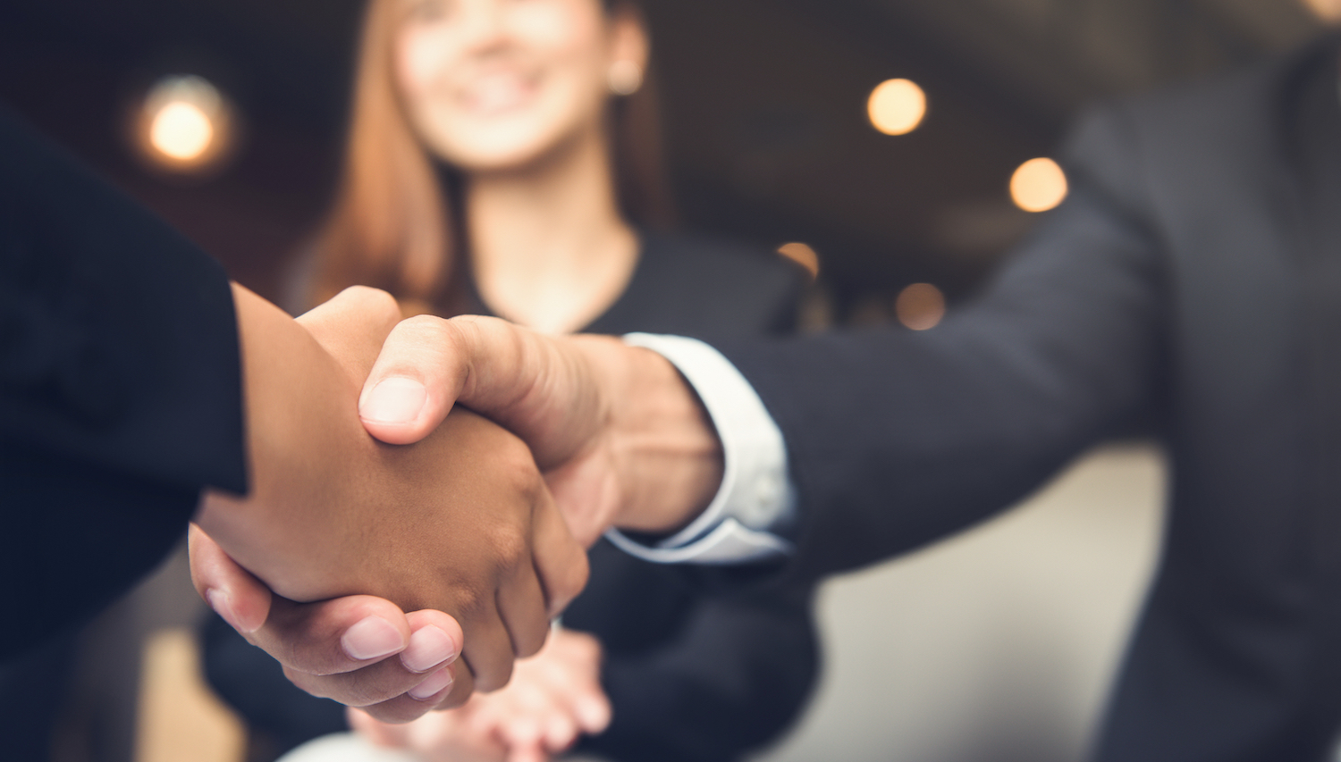 In the foreground, two hands meet in a handshake. A woman looks on in the background.