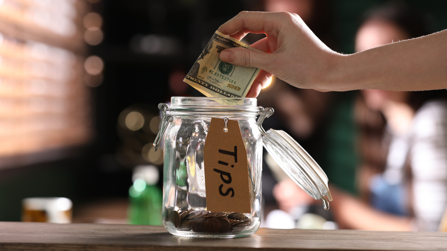 A woman's puts a $20 bill into tips into glass jar labeled "Tips" on a wooden tabletop.