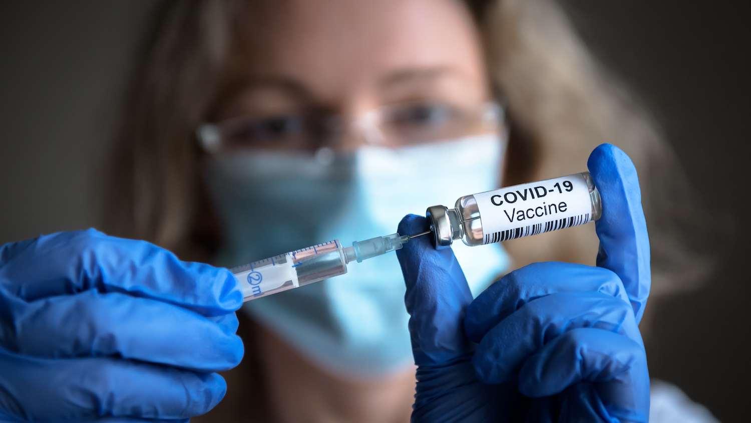 A woman wearing blue latex gloves and a blue face mask fills a syringe from a vial labeled "COVID 19 vaccine."