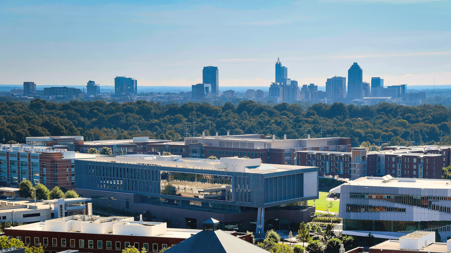 A photo highlighting the engineering buildings, particularly the Fitts-Woolard building, on Centennial Campus, in relation to downtown Raleigh skyline